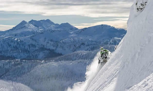 A snowmobiler climbing a steep alpine slope with mountains behind, near Revelstoke